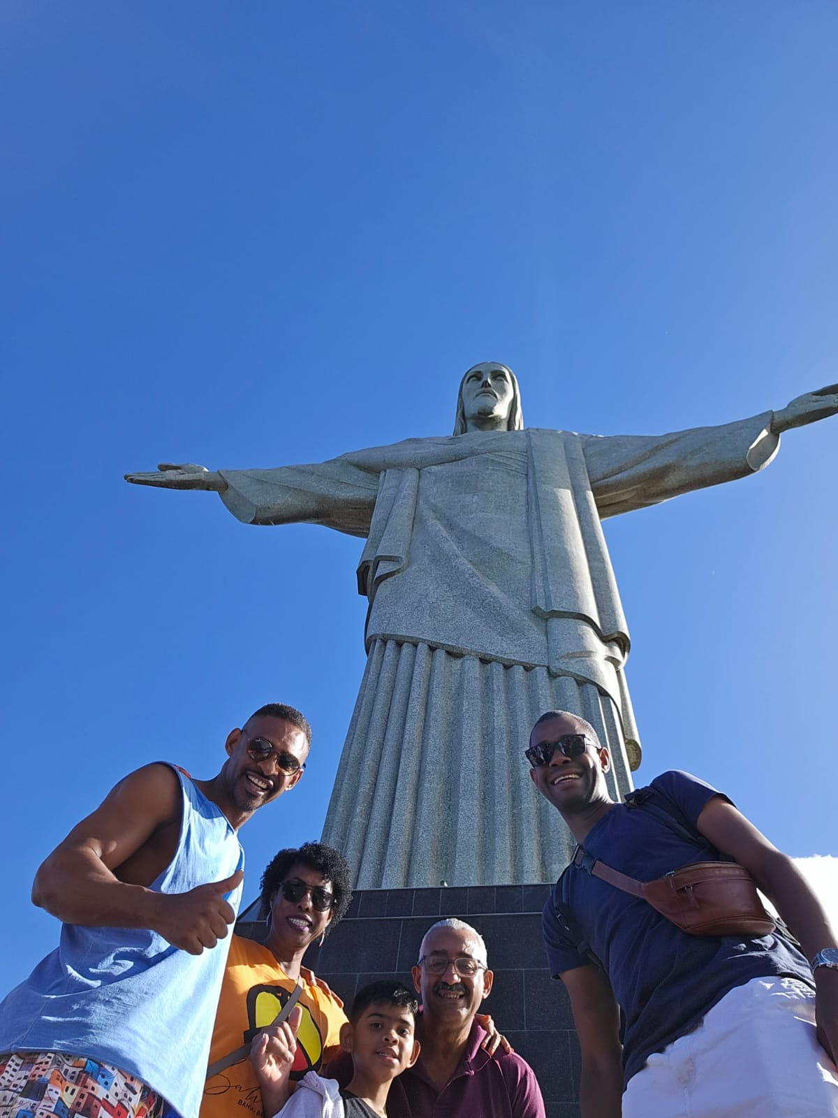 A imagem mostra um grupo de cinco pessoas posando em frente à estátua do Cristo Redentor, no Rio de Janeiro, em um dia de céu limpo e azul. A estátua monumental ocupa o centro da imagem, com seus braços estendidos. As pessoas estão posicionadas na parte inferior da foto, sorrindo e demonstrando alegria. Quatro delas são adultas e uma é uma criança. Todos estão usando óculos escuros e roupas leves, típicas de um dia ensolarado.