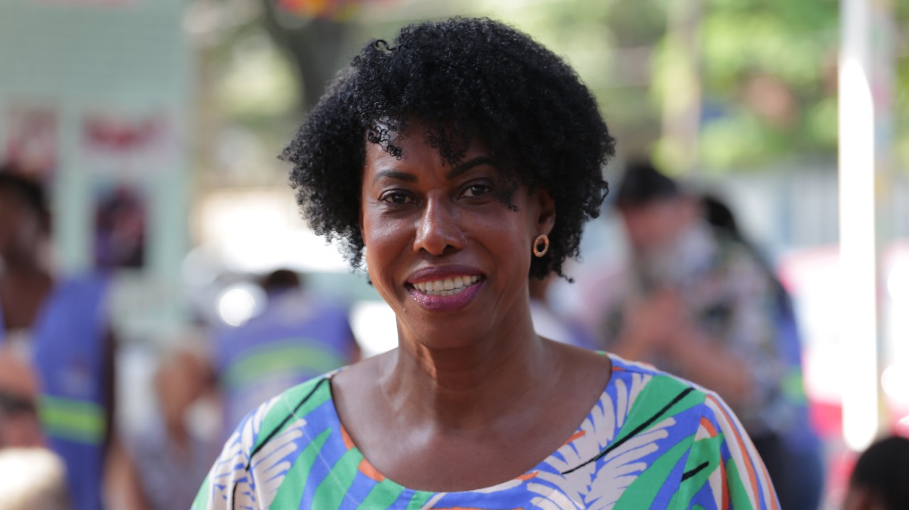 A imagem mostra Maria Isabel, uma mulher negra, com cabelo curto e cacheado, usando brincos redondos e sorrindo. Ela veste uma blusa colorida com estampas de folhas em tons de verde, azul e laranja. 