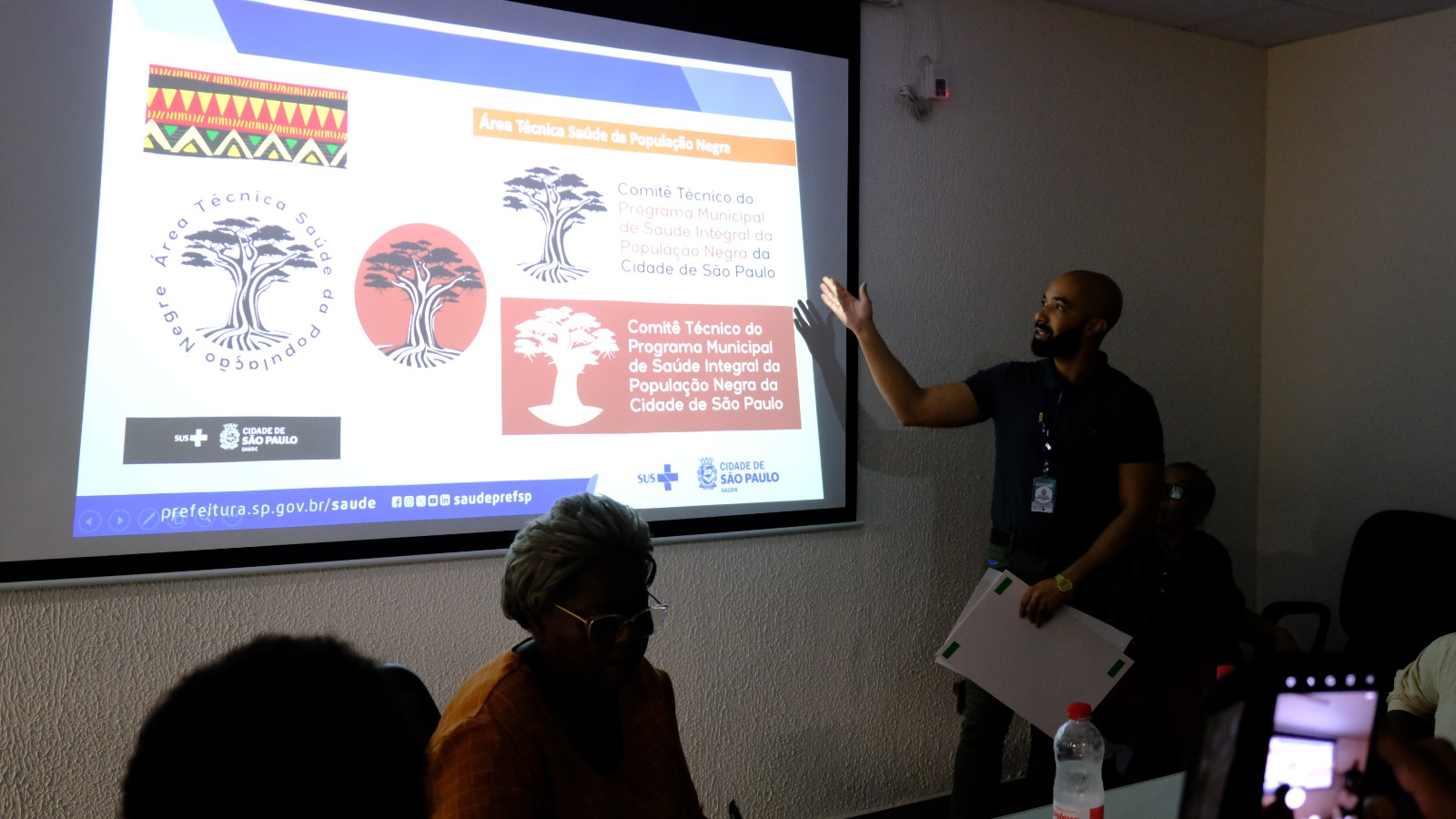 A foto mostra um homem negro e jovem falando em frente a uma projeção, na qual se vê a ilustração de uma árvore baobá e lê-se: "Comitê Técnico do Programa Municipal de Saúde Integral da População Negra da Cidade de São Paulo"