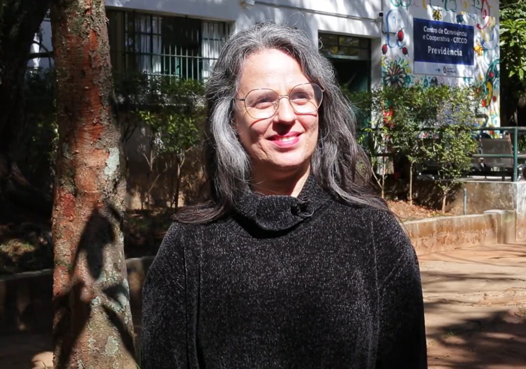 A imagem mostra uma mulher de cabelos grisalhos, com óculos e uma expressão sorridente, posando em um ambiente externo iluminado pela luz do sol. Ela veste um suéter preto com textura, e ao fundo há árvores, um muro baixo e um prédio com uma placa que indica ser o Centro de Convivência e Cooperativa - CECCO Previdência. O ambiente é rodeado por vegetação e tem uma atmosfera acolhedora, com detalhes coloridos na parede do prédio, sugerindo uma área voltada para atividades comunitárias ou sociais