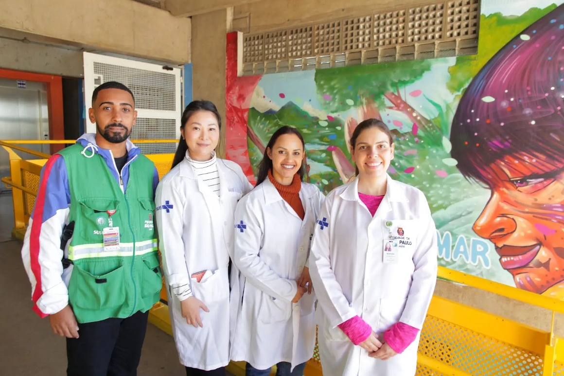 A foto mostra três mulheres e um homem uniformizados (elas de jaleco branco e ele com um colete verde) posando em frente a um mural colorido, que mostra um rosto indígena, e no qual se consegue ler a palavra "mar"