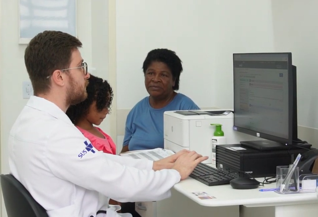 A imagem mostra uma consulta médica em um ambiente de atendimento do Sistema Único de Saúde (SUS). Um profissional de saúde, vestindo jaleco branco e óculos, está sentado em frente a um computador, interagindo com duas pacientes. Ao lado dele, uma menina com cabelos cacheados e roupa rosa está sentada, acompanhada por uma mulher mais velha, que veste uma blusa azul. No local, há equipamentos como um monitor, teclado, impressora e frascos de álcool em gel, reforçando a organização e higiene do ambiente clínico