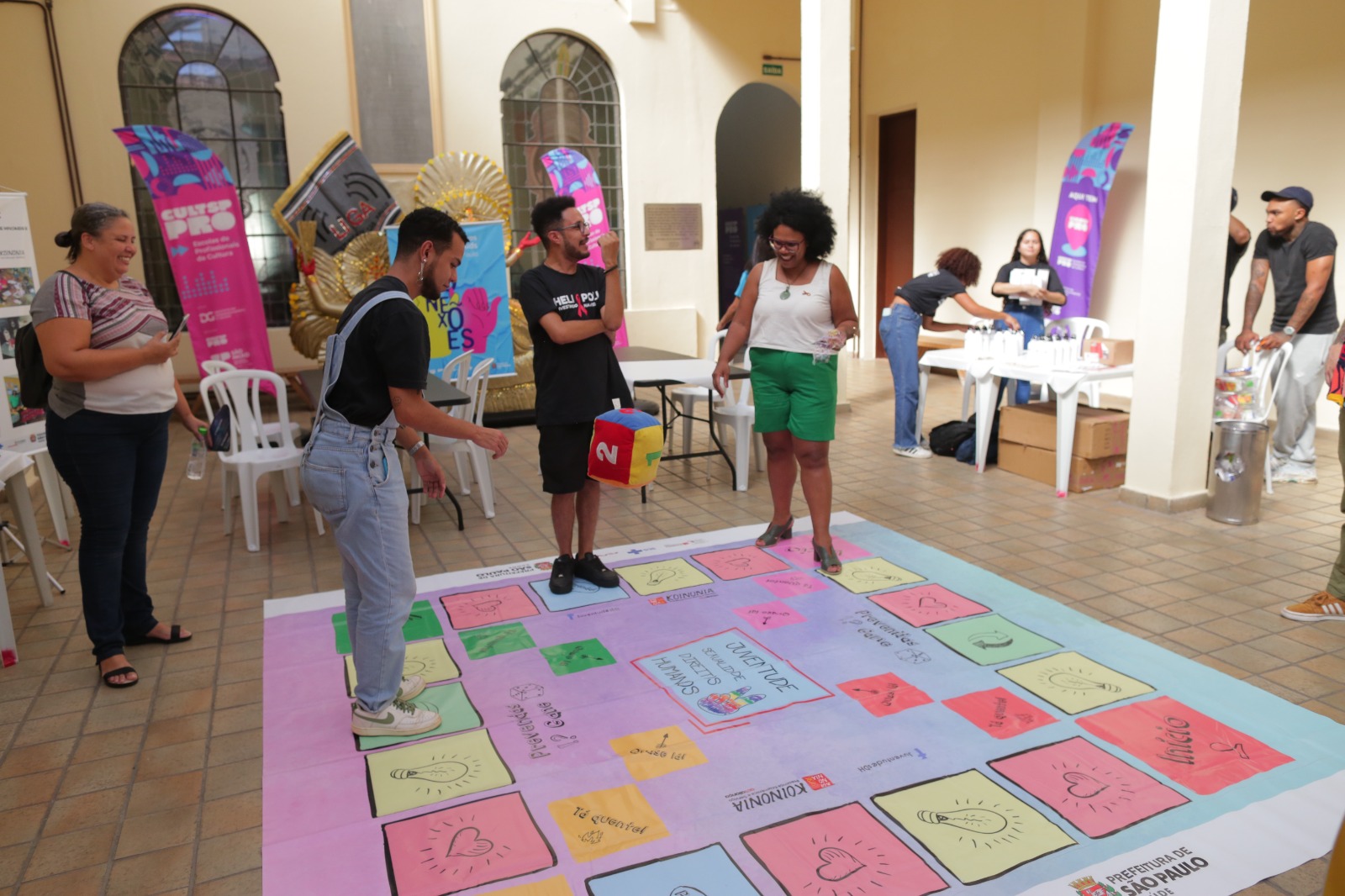 A imagem retrata um grupo de pessoas participando do jogo de tabuleiro que aborda a temática de prevenção. No centro, há um grande tabuleiro colorido no chão, com desenhos de ícones como corações, lâmpadas e mensagens. Quatro pessoas estão envolvidas no jogo: uma mulher de cabelos cacheados e blusa branca segura uma ficha, outra pessoa joga um dado colorido grande, enquanto os outros participantes observam animadamente.