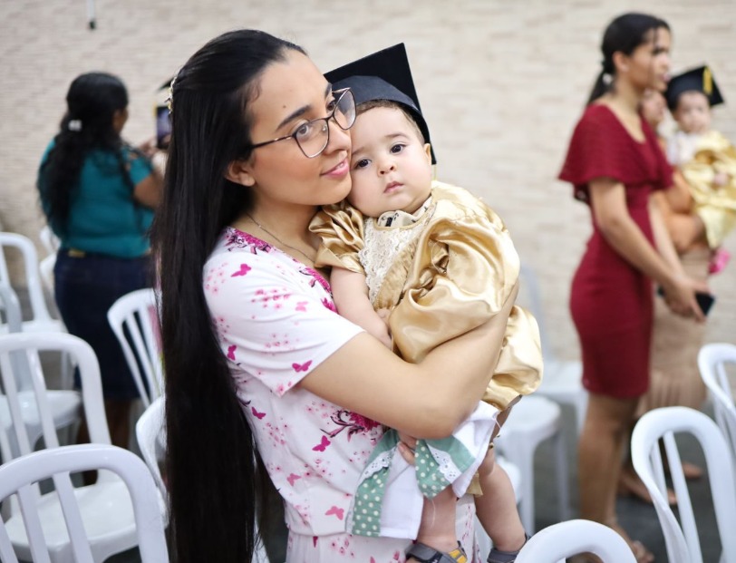 A imagem mostra uma mulher jovem com cabelos longos e óculos segurando um bebê no colo. O bebê está vestido com uma roupa dourada e usa um chapéu de formatura, indicando um evento comemorativo. A mulher olha para o bebê com carinho, enquanto ele observa a câmera com uma expressão tranquila. Ao fundo, é possível ver outras pessoas e cadeiras brancas, sugerindo que o evento ocorre em um ambiente organizado e festivo