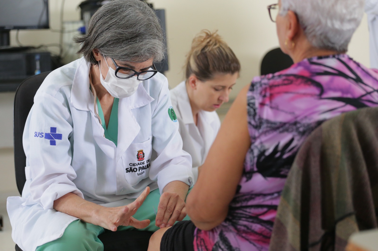 A imagem mostra a médica Simone inserirndo agulhas no joelho de uma paciente. Outra profissional aparece ao fundo.