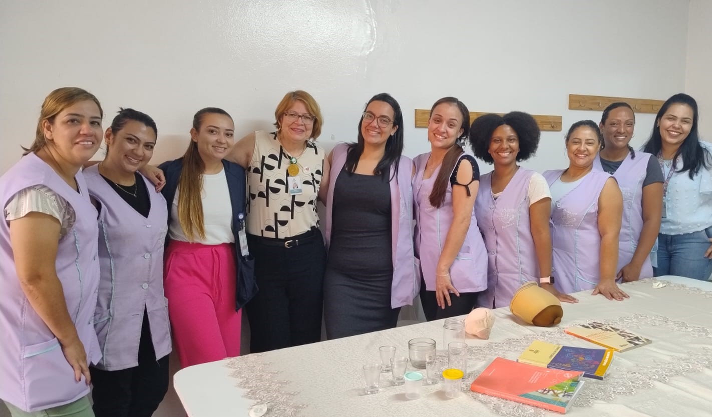 A imagem mostra um grupo de mulheres reunidas em um ambiente interno, provavelmente em uma sala de aula ou treinamento. Algumas delas vestem uniformes lilás, enquanto outras estão com roupas casuais. Todas estão sorrindo e aparentam estar participando de uma atividade conjunta. Na mesa em frente ao grupo, há objetos como copos, livros, e equipamentos que sugerem um treinamento ou atividade educacional na área de saúde ou cuidados