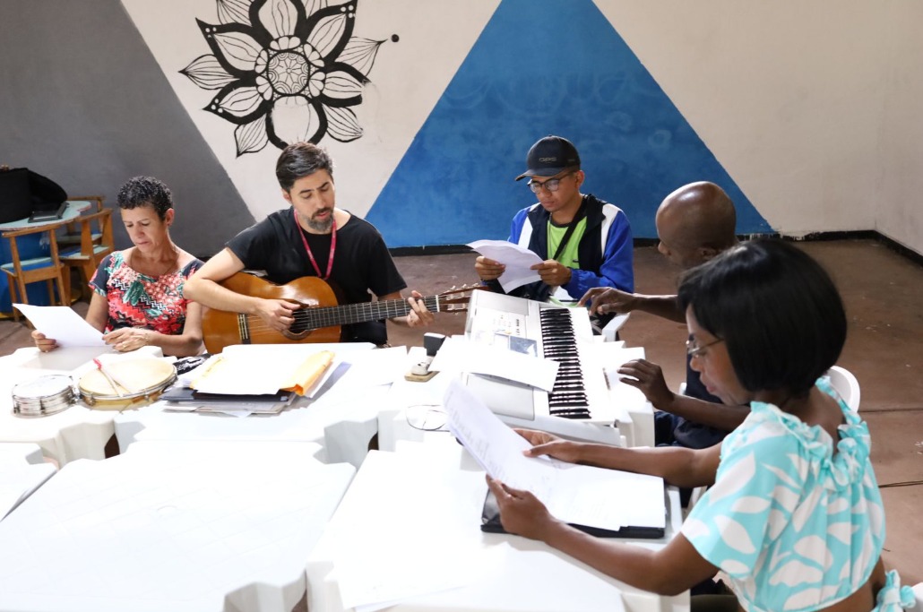 A imagem mostra um grupo de pessoas em uma atividade musical. No centro, um homem toca violão enquanto outros participantes acompanham lendo partituras ou folhas com letras de músicas. À direita, uma pessoa está tocando teclado. Na mesa, há papéis, um pandeiro e outros materiais relacionados à música. O ambiente é descontraído, com paredes decoradas, incluindo uma grande ilustração de flor em preto e branco e um triângulo azul como fundo