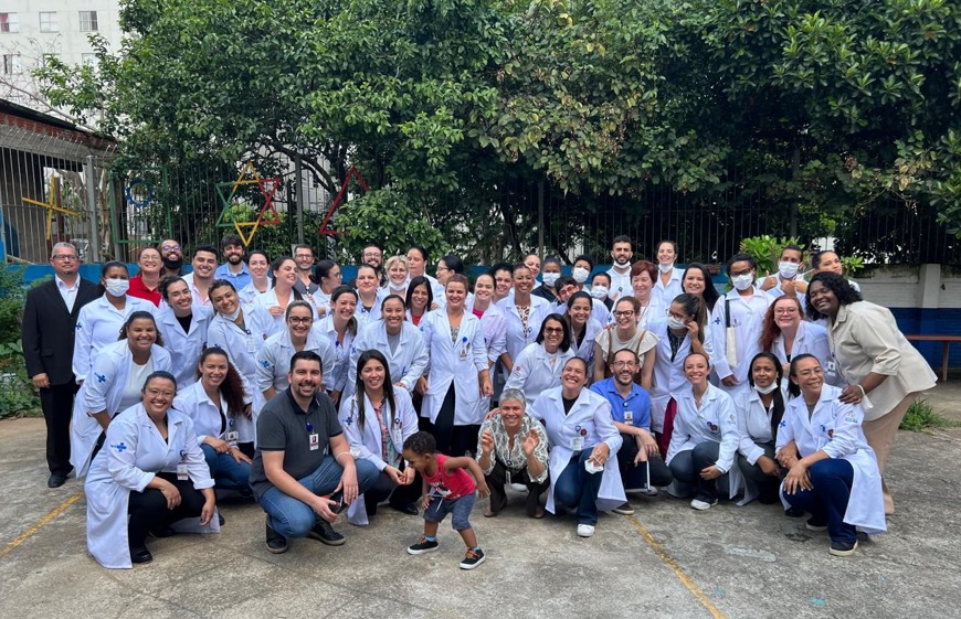 A imagem mostra um grupo grande de pessoas reunidas ao ar livre, em um ambiente arborizado, aparentemente um pátio. A maioria está usando jalecos brancos; no centro da foto, há algumas pessoas agachadas e uma criança de pé brincando, interagindo com os adultos ao redor; no fundo, há árvores, uma cerca de metal e estruturas coloridas; clima é de descontração e colaboração, com muitos sorrisos e interações entre os participantes