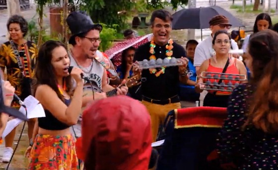 A imagem mostra um grupo de pessoas participando de uma celebração ou evento festivo ao ar livre. No centro, um homem sorridente veste uma camisa preta e calças amarelas, além de um colar colorido. Ao lado dele, uma mulher de blusa laranja e óculos. Na frente, uma jovem com saia estampada e top preto canta ao microfone, enquanto um homem ao seu lado toca um instrumento de cordas, usando um chapéu e uma camiseta sem mangas. Há outras pessoas ao fundo, algumas segurando guarda-chuvas, e a cena parece acontecer sob uma leve chuva. O ambiente é descontraído e animado.