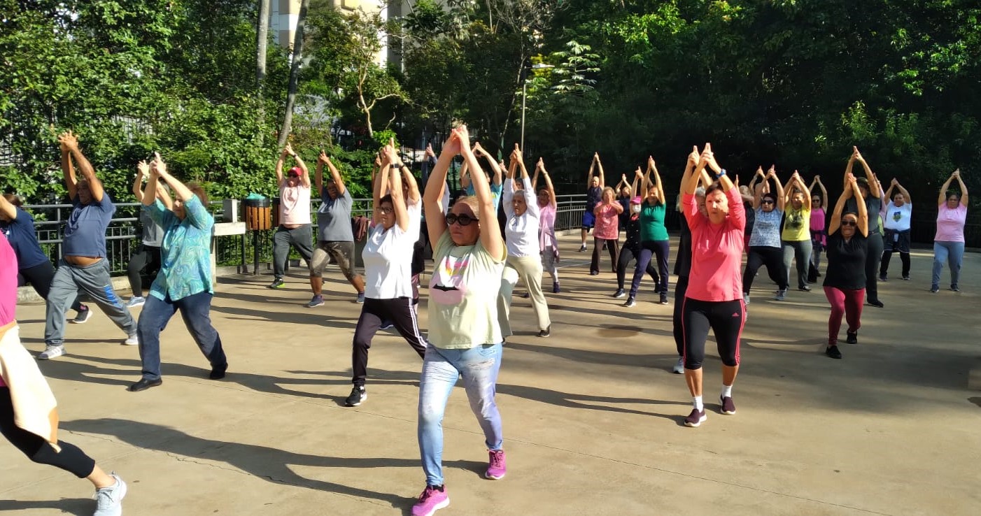 A imagem mostra um grupo grande de pessoas praticando exercícios físicos ao ar livre, em uma área pavimentada rodeada por vegetação. Os participantes, de diferentes idades — a maioria adultos e idosos —, estão com as pernas afastadas e os braços erguidos acima da cabeça, imitando o movimento de alongamento ou uma posição comum em aulas de ginástica coletiva.  O dia está ensolarado, e todos parecem estar focados na atividade. As roupas são esportivas e confortáveis, e algumas pessoas usam óculos escuros ou bonés para se proteger do sol.