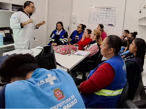 A imagem mostra uma sala de reuniões onde um grupo de profissionais da saúde está reunido. Um homem, usando jaleco branco, está de pé à frente do grupo, aparentemente ministrando uma capacitação ou treinamento. Os participantes estão sentados ao redor de uma mesa, atentos à apresentação. Muitos deles vestem coletes azuis com o logotipo do SUS (Sistema Único de Saúde) e da Prefeitura de São Paulo