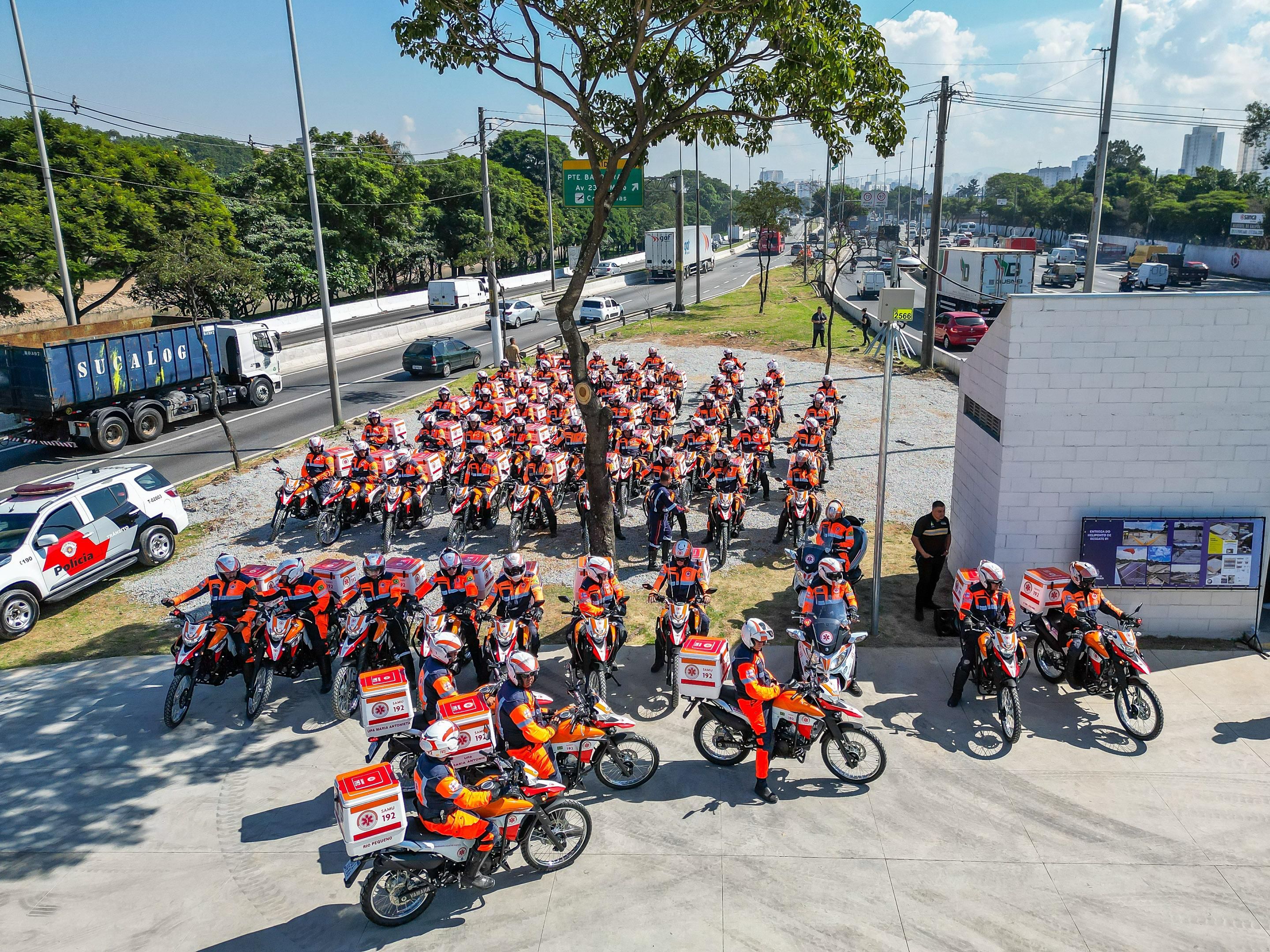 A imagem mostra uma vista aérea de uma concentração organizada de profissionais do SAMU (Serviço de Atendimento Móvel de Urgência) com motocicletas. Há dezenas de motos perfiladas, estacionadas em fileiras ordenadas, todas equipadas com caixas de emergência nas cores branca, laranja e vermelha, contendo o número "192" e a identificação do serviço. Os profissionais vestem uniformes de alta visibilidade na cor laranja com azul escuro, além de capacetes nas cores branca, azul e laranja.  Ao redor da área onde estão concentradas as motos, é possível observar uma movimentada rodovia urbana, com diversos veículos trafegando. À esquerda, há um carro da polícia estacionado. O local possui árvores, postes de iluminação e placas indicando uma área urbana. Ao fundo, vê-se uma linha de prédios ao longe e o céu parcialmente nublado.
