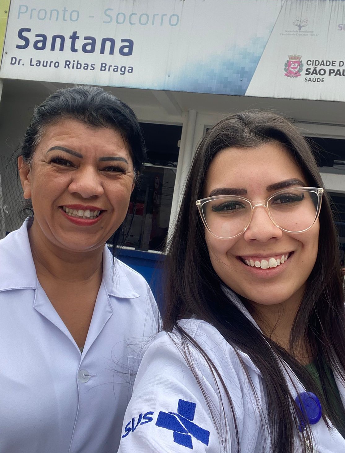 A imagem mostra duas profissionais de saúde sorrindo, ambas vestidas com jalecos brancos do SUS (Sistema Único de Saúde), em frente à entrada do Pronto-Socorro Santana – Dr. Lauro Ribas Braga, unidade da Prefeitura de São Paulo. Ao fundo, é possível ver a placa de identificação do equipamento de saúde com os logotipos oficiais da prefeitura e da Secretaria Municipal da Saúde. A foto transmite um clima de acolhimento e profissionalismo, destacando o trabalho das equipes de enfermagem na rede pública