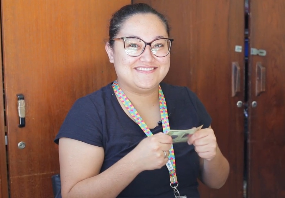 A imagem mostra uma mulher sorridente segurando um documento de identidade. Ela está usando óculos, veste uma camiseta preta e porta um cordão colorido com estampa geométrica no pescoço, característico de pessoas com transtorno do espectro autista