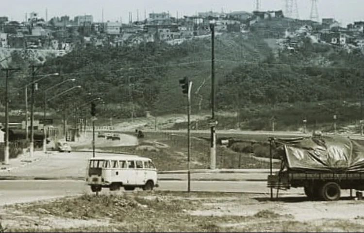 Vale do Rio Aricanduva no ano de 1970 com uma Kombi parada no semáforo e a parte de trás de uma caçamba de caminhão coberto com lona, no horizonte a avenida aricanduva e de fundo um morro com árvores e algumas casas.