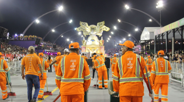 Uma foto noturna e panorâmica captura um momento vibrante do carnaval. Em primeiro plano, um grupo de garis, vestindo uniformes laranja com faixas refletivas e bonés da mesma cor, está de costas para a câmera, varrendo a rua. Eles utilizam vassouras com cabos de madeira e cerdas marrons e vermelhas.  Ao fundo, uma alegoria imponente e iluminada em tons de branco e dourado se destaca. A alegoria parece representar uma figura mitológica ou um brasão com uma águia de asas abertas no topo. A rua está cercada por arquibancadas lotadas de espectadores, criando uma atmosfera festiva e animada.  A iluminação artificial é intensa, com diversos postes de luz arqueados sobre a rua, projetando uma luz branca e brilhante sobre a cena. O céu noturno serve de pano de fundo para essa celebração cultural. 