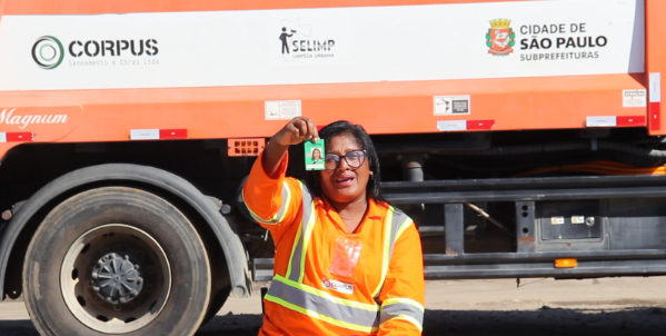Uma mulher de pele morena, com óculos de armação escura e cabelos escuros presos, está no centro da imagem, sorrindo e olhando para a câmera. Ela veste um uniforme laranja com faixas refletivas cinzas e amarelas. Sua mão direita está levantada, segurando um crachá verde com uma foto colorida de seu rosto.  Ao fundo, parcialmente visível, está um caminhão de cor laranja com detalhes em branco e vermelho. Na lateral do caminhão, pode-se ler as palavras "CORPUS" em letras verdes dentro de um círculo, "SELIMP" com um pictograma de uma pessoa varrendo, e um logotipo com a inscrição "CIDADE DE SÃO PAULO SUBPREFEITURAS" e um desenho que inclui o brasão da cidade. A roda traseira do caminhão é preta e grande. O chão é de asfalto. 