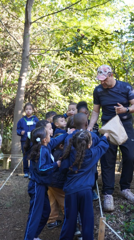  A fotografia mostra um grupo de crianças pequenas, vestindo uniformes azuis com faixas brancas ou claras, reunidas em torno de um homem adulto em um ambiente arborizado. O homem, que parece ser um educador ou guia, usa uma camiseta preta, calças escuras, um boné estampado e tênis. Ele segura uma sacola de papel em uma das mãos, enquanto as crianças o observam atentamente, algumas esticando as mãos em sua direção.  As crianças têm diferentes tons de pele e texturas de cabelo, algumas com cabelos escuros e lisos, outras com cachos ou tranças. Ao fundo, árvores e vegetação densa sugerem um local ao ar livre, como um parque, floresta ou jardim botânico. Há uma corda esticada e fixada em estacas baixas no chão, delimitando uma área. A luz do sol ilumina a cena, criando um contraste entre a sombra e a luz.