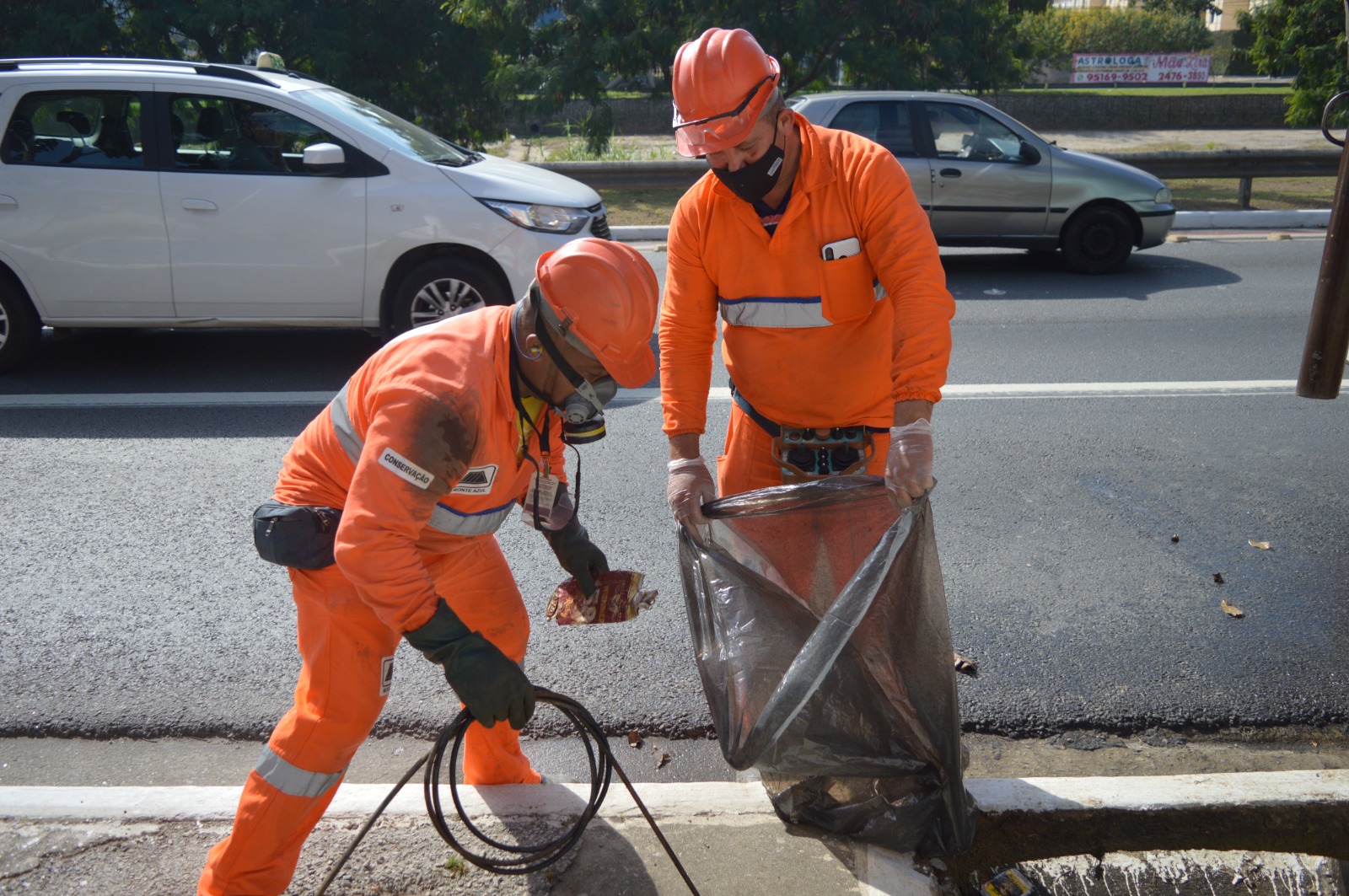 A foto mostra dois funcionários ao centro da imagem, executando a limpeza com hidrojato. Um deles está meio inclinado usando equipamento de epis, e outro está de pé, segurando um saco plástico.