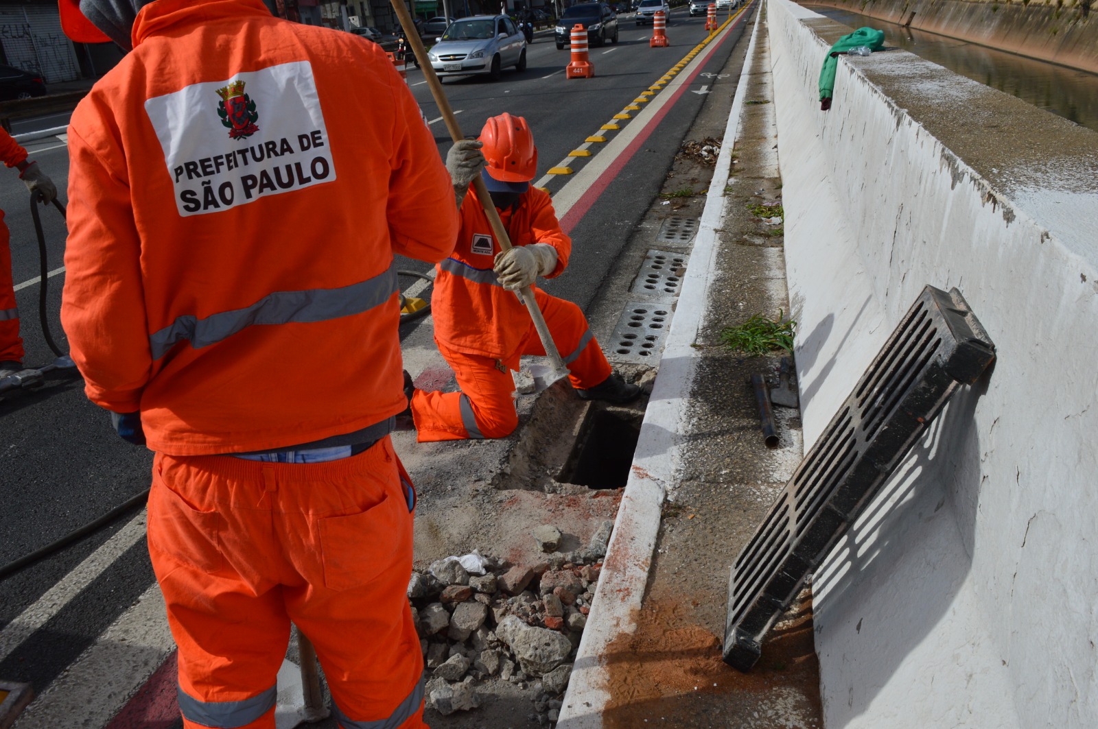 A foto mostra dois funcionários executando a limpeza manual de bocas de lobo em uma rua. Um funcionário aparece de costas em pé, e o logo da Prefeitura de São Paulo está em destaque no uniforme. Outro funcionário está com um joelho no chão, enquanto executa a limpeza.