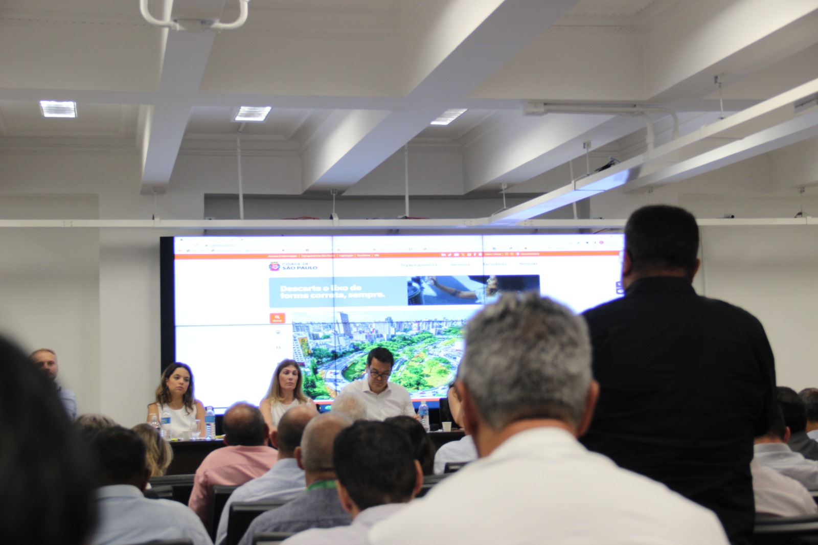 A foto tirada ao fundo do auditório mostra a mesa principal em foco, enquanto um fotografado de costas usando camisa social preta fala ao microfone. 