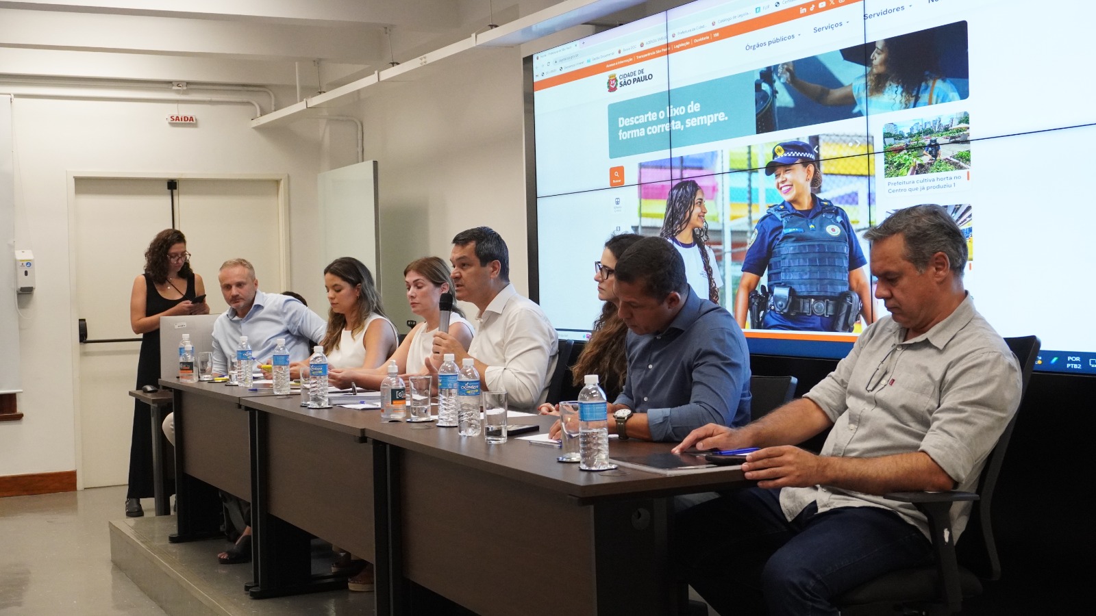 A foto tirada da mesa composta do auditório, mostra sete pessoas sentadas. Uma mulher de vestido preto para do lado esquerdo da foto de pé, usando o celular. Em plano principal está um homem de camisa social branca falando no microfone. 