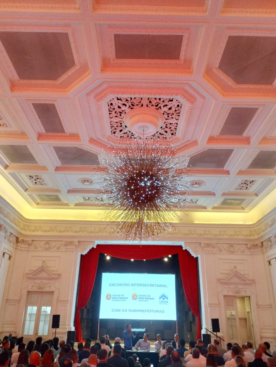 A foto mostra um grande salão com um lustre moderno e brilhante pendurado no teto ornamentado com detalhes clássicos em tons claros. Ao fundo, um palco com cortinas vermelhas está montado para um evento chamado "ENCONTRO INTERSECRETARIAL", conforme escrito em uma tela iluminada. Abaixo do título, há logotipos da "CIDADE DE SÃO PAULO" e "COHAB". No palco, um homem está de pé, falando para uma plateia de diversas pessoas que estão sentadas em cadeiras, olhando em direção ao palco. A iluminação ambiente é suave, criando um clima formal e de evento.
